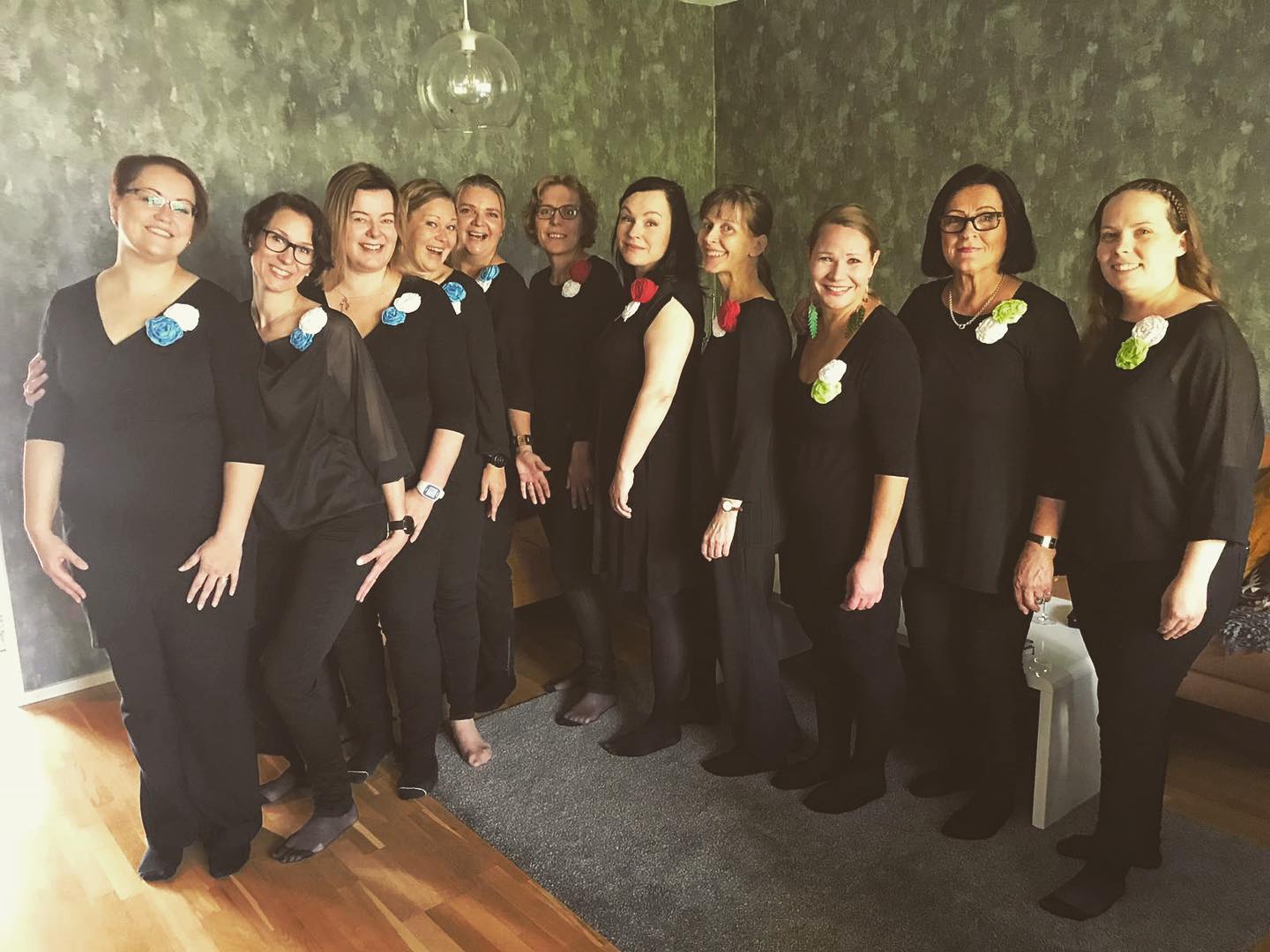 9 kuorolaulajanaista huoneessa, jossa vihreä tapetti. Naiset ovat menossa laulukeikalle. 